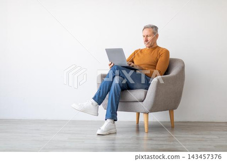 Focused Elderly Caucasian Man Working On Laptop While Sitting Comfortably In A Cozy Chair At Home. Engaged In Work Or Personal Project, Creating A Productive Environment. Business Concept. Focused Elderly Caucasian Man Working On Laptop While Sitting Comfortably In A Cozy Chair At Home. Engaged In Work Or Personal Project, Creating A Productive Environment. Business Concept. 143457376