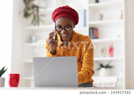 Smiling Black Woman Using Laptop And Holding Phone While Engaging In Work, Sitting At Home Office Desk. Focused And Productive Environment, Business Concept. Smiling Black Woman Using Laptop And Holding Phone While Engaging In Work, Sitting At Home Office Desk. Focused And Productive Environment, Business Concept. 143457392