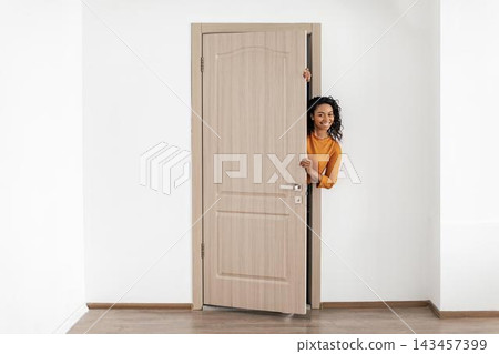 Happy Black Woman Smiling While Peeking Through Doorway In Modern Interior. Showing Joy And Positivity In A Friendly Home Ambiance. Concept Of Happiness And Welcoming Atmosphere. Happy Black Woman Smiling While Peeking Through Doorway In Modern Interior. Showing Joy And Positivity In A Friendly Home Ambiance. Concept Of Happiness And Welcoming Atmosphere. 143457399