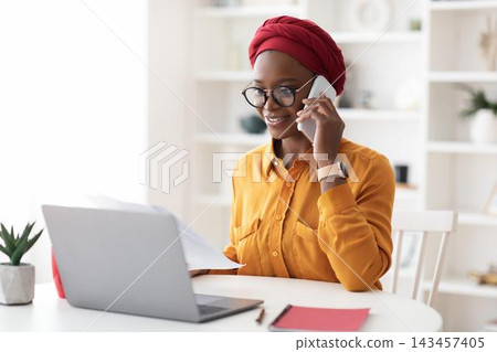 Smiling Young Black Woman Engaged In Phone Conversation While Sitting At Home Office Desk, Using Laptop To Work On Documents And Organize Tasks, Modern Workspace. Business Concept. Smiling Young Black Woman Engaged In Phone Conversation While Sitting At Home Office Desk, Using Laptop To Work On Documents And Organize Tasks, Modern Workspace. Business Concept. 143457405