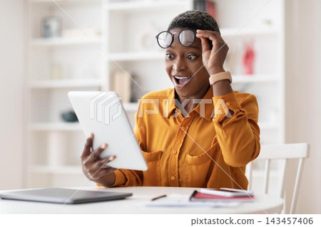 Excited Black Female With Short Hair, Expressing Joy While Using Tablet At Home, Sitting At Kitchen Table. Engaging In Digital Activity With A Happy Smile, Relaxed Atmosphere. Excited Black Female With Short Hair, Expressing Joy While Using Tablet At Home, Sitting At Kitchen Table. Engaging In Digital Activity With A Happy Smile, Relaxed Atmosphere. 143457406