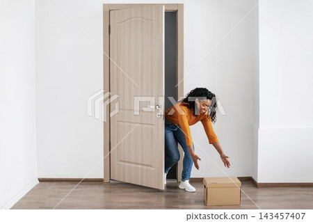 Excited Happy Black Woman Opening Door To Receive A Package, Kneeling To Grab Box In Bright Modern Interior Of Home. Cheerful Adult Female Enjoying Unboxing Experience At Home, Lifestyle Concept. Excited Happy Black Woman Opening Door To Receive A Package, Kneeling To Grab Box In Bright Modern Interior Of Home. Cheerful Adult Female Enjoying Unboxing Experience At Home, Lifestyle Concept. 143457407