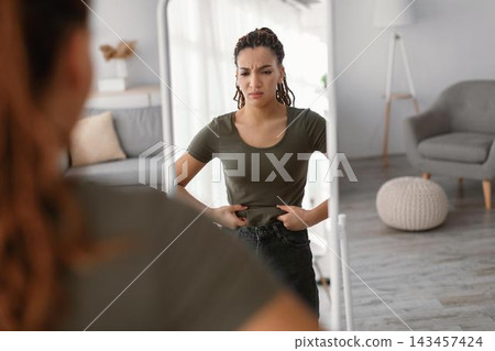 Worried Young Black Woman Staring At Reflection In Mirror While Adjusting Clothing, Showing Discontent With Appearance, In Modern Living Room. Acknowledging Body Image And Weight Loss Concept. Worried Young Black Woman Staring At Reflection In Mirror While Adjusting Clothing, Showing Discontent With Appearance, In Modern Living Room. Acknowledging Body Image And Weight Loss Concept. 143457424