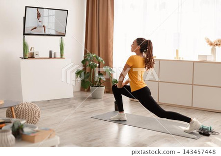 Focused Young Black Woman Engaged In Home Workout Routine Following Online Tutorial, Performing Lunges On Mat In Bright Living Room, Exploring Fitness And Health From Home. Focused Young Black Woman Engaged In Home Workout Routine Following Online Tutorial, Performing Lunges On Mat In Bright Living Room, Exploring Fitness And Health From Home. 143457448