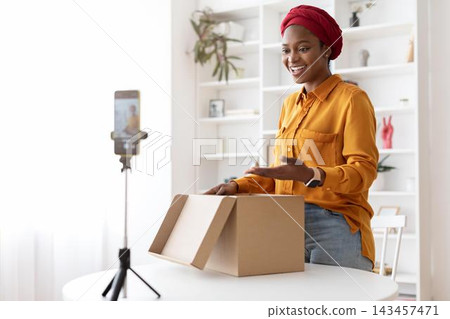 Smiling Black Woman Filming Unboxing Video While Opening A Package At Home, Showing Excitement And Joy. Engaged In Content Creation In A Bright Room With Modern Decor, Business Concept. Smiling Black Woman Filming Unboxing Video While Opening A Package At Home, Showing Excitement And Joy. Engaged In Content Creation In A Bright Room With Modern Decor, Business Concept. 143457471