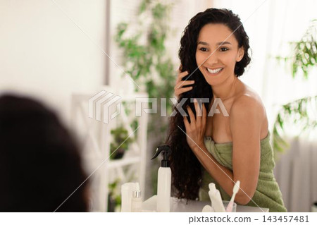 Smiling Young Woman With Curly Hair Applying Hair Product In Front Of Mirror At Home, Taking Care Of Her Hair And Wellness. Home Setting, Beauty And Self-care Concept. Smiling Young Woman With Curly Hair Applying Hair Product In Front Of Mirror At Home, Taking Care Of Her Hair And Wellness. Home Setting, Beauty And Self-care Concept. 143457481