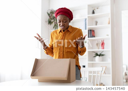 Worried Expression On Black Woman In Late 20s, Unboxing Empty Brown Package At Office, Confused Reaction, Emotion Displayed While In Modern Workspace, Relevant To Delivery Concept. Worried Expression On Black Woman In Late 20s, Unboxing Empty Brown Package At Office, Confused Reaction, Emotion Displayed While In Modern Workspace, Relevant To Delivery Concept. 143457540