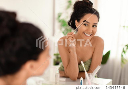Smiling Young Woman With Natural Beauty Holding Eyebrow Brush While Looking At Mirror In Cozy Bathroom. Engaged In Self-care Routine, Enjoying Skincare At Home, Beauty Concept. Smiling Young Woman With Natural Beauty Holding Eyebrow Brush While Looking At Mirror In Cozy Bathroom. Engaged In Self-care Routine, Enjoying Skincare At Home, Beauty Concept. 143457546
