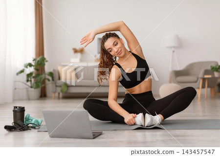 Focused Young Woman Stretching While Sitting On Yoga Mat At Home, Engaging In Exercise Routine For Fitness And Wellness In Modern Living Room Setting. Weight Loss Concept. 143457547
