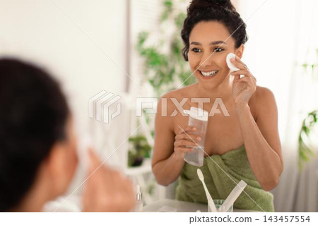 Smiling Young Woman Applying Skincare Product With Cotton Pad In Front Of A Mirror At Home, Taking Care Of Her Skin With A Routine, Emphasizing Personal Wellness And Relaxation Concept. Smiling Young Woman Applying Skincare Product With Cotton Pad In Front Of A Mirror At Home, Taking Care Of Her Skin With A Routine, Emphasizing Personal Wellness And Relaxation Concept. 143457554