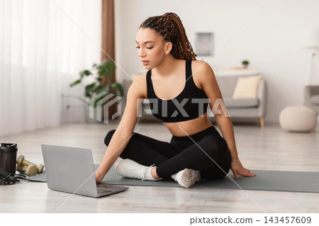 Focused Young Black Woman Sitting Cross-legged On Exercise Mat, Engaging In Online Fitness Routine At Home, Using Laptop, Promoting Healthy Lifestyle And Wellbeing. Fitness Concept. 143457609