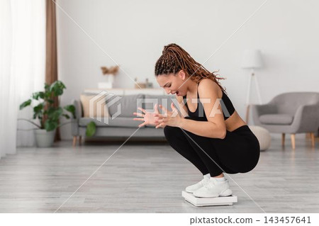 Worried Black Woman In Late 20s Weighing Herself While Squatting Down On Scale At Home With Modern Interior. Focus On Weight Measurement And Fitness Journey. Weight Loss Concept. 143457641
