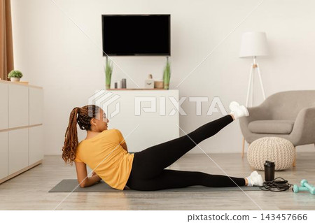 Focused Black Female Exercising At Home On Yoga Mat, Doing Side Leg Raises In Modern Living Room. Staying Active And Fit With Home Workouts, Emphasizing Importance Of Fitness Concept. 143457666