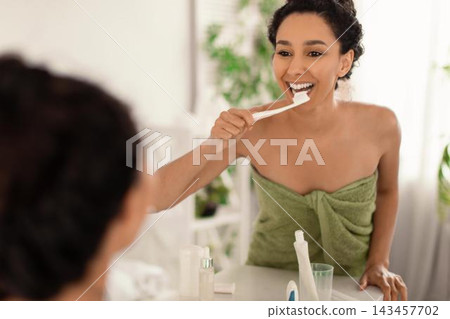 Smiling Young African American Woman Brushing Her Teeth In Front Of Mirror While Enjoying Morning Routine At Home In Bathroom. Personal Care And Skincare Concept. 143457702