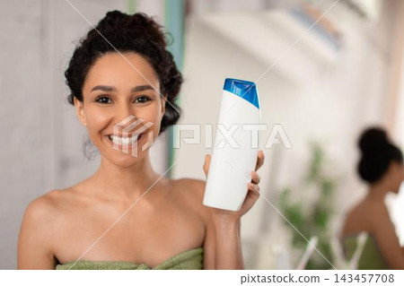 Smiling Young Middle Eastern Woman Holding A Shampoo Bottle In Bathroom, Happy And Cheerful While Showing Hair Care Product, Standing By The Mirror, Home Skincare Concept. 143457708