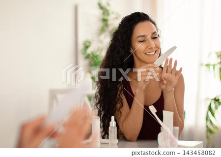 Smiling Young Hispanic Woman Focused On Grooming Her Nails With A File, Sitting At Home Setting With Skincare Products On The Table. Engaged And Happy In Her Personal Care Routine. Smiling Young Hispanic Woman Focused On Grooming Her Nails With A File, Sitting At Home Setting With Skincare Products On The Table. Engaged And Happy In Her Personal Care Routine. 143458082