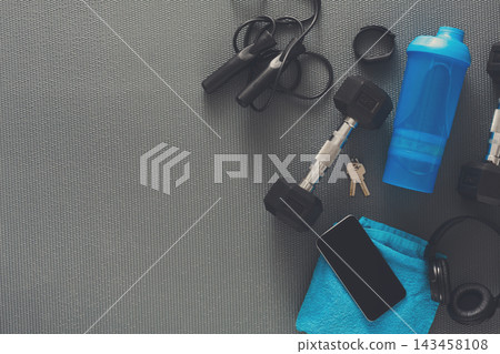 Two dumbbells are placed on a grey mat alongside a water bottle, a towel, a phone, and workout gear such as skipping ropes and headphones. Keys are also present in the scene. Two dumbbells are placed on a grey mat alongside a water bottle, a towel, a phone, and workout gear such as skipping ropes and headphones. Keys are also present in the scene. 143458108