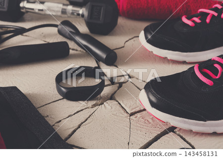 A set of workout shoes, weights, and a fitness tracker are arranged on a cracked floor. A jump rope and a towel are also present, indicating preparation for exercise at home. A set of workout shoes, weights, and a fitness tracker are arranged on a cracked floor. A jump rope and a towel are also present, indicating preparation for exercise at home. 143458131