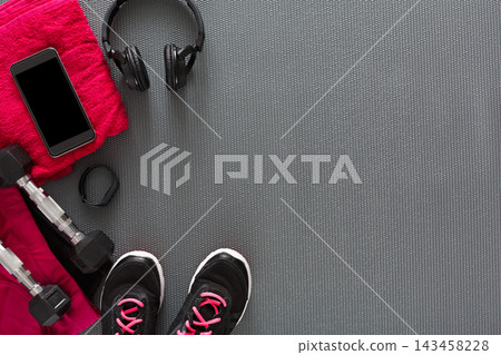A workout setup on a gym floor includes dumbbells, a smartphone, a towel, headphones, and athletic shoes. The items are arranged neatly and ready for a fitness session. A workout setup on a gym floor includes dumbbells, a smartphone, a towel, headphones, and athletic shoes. The items are arranged neatly and ready for a fitness session. 143458228