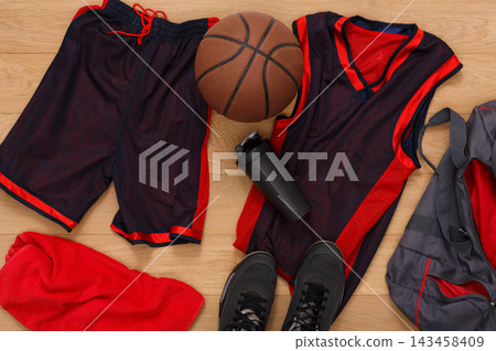 Basketball uniform, red towel, basketball, water bottle, and shoes are arranged on a wooden floor. The items are ready for a practice session or game in a gym environment. Basketball uniform, red towel, basketball, water bottle, and shoes are arranged on a wooden floor. The items are ready for a practice session or game in a gym environment. 143458409