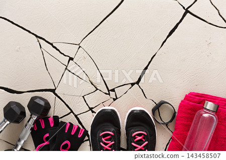 Workout gear is laid on a cracked floor. The items include dumbbells, a water bottle, gloves, a towel, and athletic shoes, creating a fitness scene ready for exercise. Workout gear is laid on a cracked floor. The items include dumbbells, a water bottle, gloves, a towel, and athletic shoes, creating a fitness scene ready for exercise. 143458507