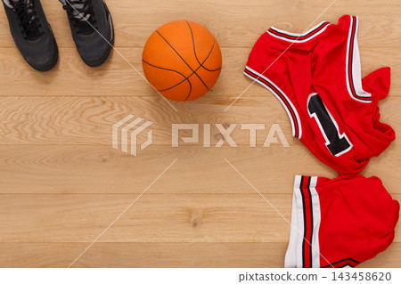 A basketball ball sits on a wooden floor next to a pair of shoes and a red jersey with number one. Red shorts are also nearby, waiting for the next game. 143458620