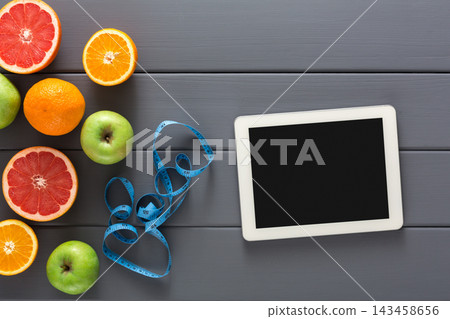 A variety of fresh fruits including oranges, apples, and grapefruits is placed on a wooden table. A measuring tape lies beside a tablet, suggesting a focus on health and diet. 143458656