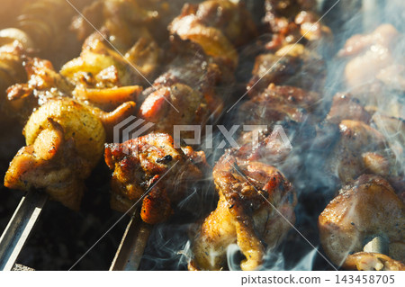 Chicken pieces are on metal skewers and cooking over an open flame. Smoke rises from the grill while the food gets hot. Friends gather around the grill enjoying the summer weather. 143458705