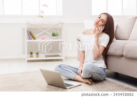 A young woman is sitting on the floor in a living room. She is talking on the phone and using a laptop in front of her. There are plants and a shelf in the background. A young woman is sitting on the floor in a living room. She is talking on the phone and using a laptop in front of her. There are plants and a shelf in the background. 143458882