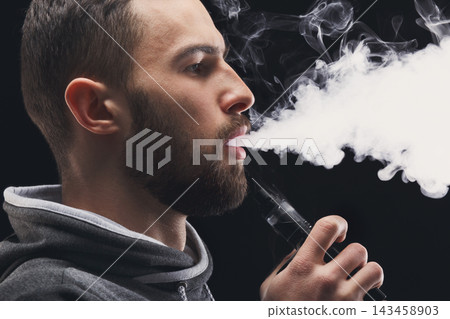 A man stands in a dark setting while using a vaping device. He exhales a large cloud of vapor. The scene shows a focus on his profile, highlighting the vapor he produces. A man stands in a dark setting while using a vaping device. He exhales a large cloud of vapor. The scene shows a focus on his profile, highlighting the vapor he produces. 143458903