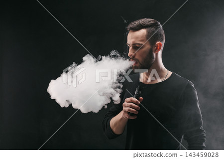 A man stands in a dark space and takes a deep puff from a vape device. He exhales a large cloud of vapor, creating a thick mist around him. The setting focuses on the vapor and his action. A man stands in a dark space and takes a deep puff from a vape device. He exhales a large cloud of vapor, creating a thick mist around him. The setting focuses on the vapor and his action. 143459028