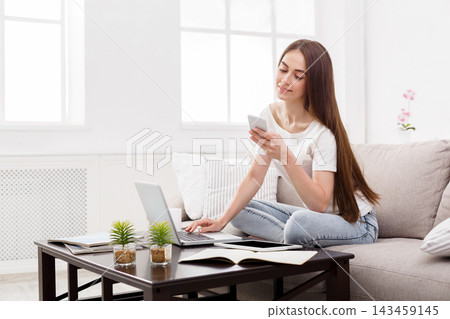 A young woman sits on a couch in a bright living room. She uses a laptop and looks at her smartphone. Small plants decorate the table next to her. A young woman sits on a couch in a bright living room. She uses a laptop and looks at her smartphone. Small plants decorate the table next to her. 143459145
