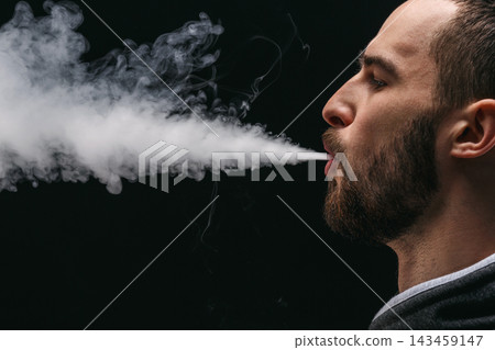 A man stands in a dark area and exhales vapor from his mouth. The vapor forms a cloud in the air. The lighting highlights his profile. The scene captures the moment of vaping. A man stands in a dark area and exhales vapor from his mouth. The vapor forms a cloud in the air. The lighting highlights his profile. The scene captures the moment of vaping. 143459147