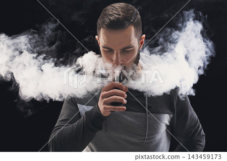 A man stands indoors, holding a vaping device. He exhales a large cloud of vapor that fills the space around him. The setting is dark, emphasizing the vapor. A man stands indoors, holding a vaping device. He exhales a large cloud of vapor that fills the space around him. The setting is dark, emphasizing the vapor. 143459173