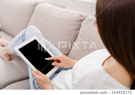 A woman sits on a couch in a bright room and uses a tablet. She points at the screen with one finger while resting her other arm. The room has simple decor and natural light. A woman sits on a couch in a bright room and uses a tablet. She points at the screen with one finger while resting her other arm. The room has simple decor and natural light. 143459175