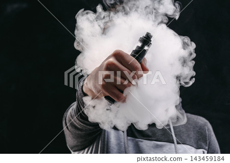 A person holds a vape device with one hand and exhales a thick cloud of vapor. The background is dark, highlighting the vapor. This scene captures the act of vaping. A person holds a vape device with one hand and exhales a thick cloud of vapor. The background is dark, highlighting the vapor. This scene captures the act of vaping. 143459184
