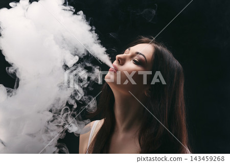 A woman stands in a dark space and releases smoke from her mouth. She is looking to the side with long hair and a calm expression while engaging in the act of vaping. A woman stands in a dark space and releases smoke from her mouth. She is looking to the side with long hair and a calm expression while engaging in the act of vaping. 143459268
