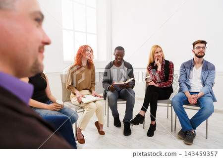 A group of six people sits in a circle in a well-lit room. They are engaged in a discussion, with one person reading from a book. Others listen and respond. 143459357