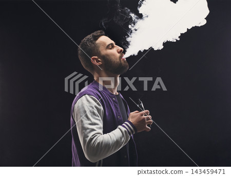 A man stands in a dark space, breathing out vapor from a vaping device. He holds the device in one hand while focusing on the thick smoke. The atmosphere suggests a casual vibe. A man stands in a dark space, breathing out vapor from a vaping device. He holds the device in one hand while focusing on the thick smoke. The atmosphere suggests a casual vibe. 143459471