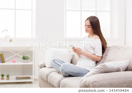 A young woman is seated comfortably on a couch in a light living room. She looks at her smartphone while surrounded by soft cushions and natural light coming through the windows. A young woman is seated comfortably on a couch in a light living room. She looks at her smartphone while surrounded by soft cushions and natural light coming through the windows. 143459484
