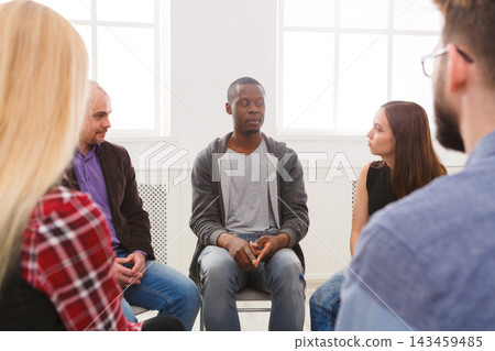 People sit in a circle in a light room. A man talks while others listen. The setting is casual and focused on sharing thoughts and experiences. People sit in a circle in a light room. A man talks while others listen. The setting is casual and focused on sharing thoughts and experiences. 143459485