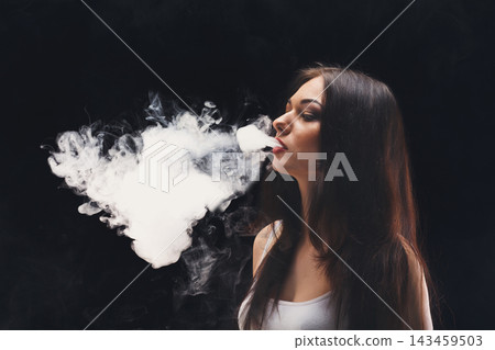 A young woman stands in a dark room. She exhales a cloud of vapor from her mouth. The vapor creates a shape in the air. Her long hair flows down past her shoulders. A young woman stands in a dark room. She exhales a cloud of vapor from her mouth. The vapor creates a shape in the air. Her long hair flows down past her shoulders. 143459503