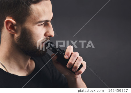 A man is using a black vaporizer indoors. He holds the device close to his mouth, focusing on the action of vaping. The background is dark, emphasizing his profile and gesture. A man is using a black vaporizer indoors. He holds the device close to his mouth, focusing on the action of vaping. The background is dark, emphasizing his profile and gesture. 143459527