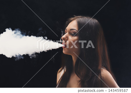 A woman stands in a dark space, exhaling a large cloud of smoke. She maintains a focused expression while releasing the vapor. This scene takes place in the evening at an indoor location. A woman stands in a dark space, exhaling a large cloud of smoke. She maintains a focused expression while releasing the vapor. This scene takes place in the evening at an indoor location. 143459587