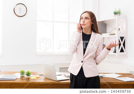 A woman stands in an office setting. She is on the phone, looking concerned, with papers and a laptop in front of her. The room has large windows that let in natural light. A woman stands in an office setting. She is on the phone, looking concerned, with papers and a laptop in front of her. The room has large windows that let in natural light. 143459730