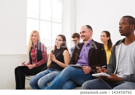 A group of diverse individuals sits in a training session. They listen closely and take notes. The venue has large windows letting in natural light, creating a welcoming environment. A group of diverse individuals sits in a training session. They listen closely and take notes. The venue has large windows letting in natural light, creating a welcoming environment. 143459792