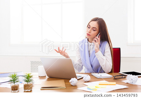 A woman is talking on her phone in an office setting. She sits at a desk with a laptop open in front of her. Papers and crumpled notes are scattered on the desk. Green plants add to the workspace. A woman is talking on her phone in an office setting. She sits at a desk with a laptop open in front of her. Papers and crumpled notes are scattered on the desk. Green plants add to the workspace. 143459793
