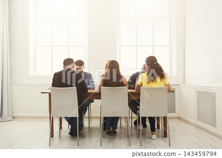A group of six individuals sits around a wooden table in a well-lit room. They appear engaged in a conversation, focusing on task-related discussions. A group of six individuals sits around a wooden table in a well-lit room. They appear engaged in a conversation, focusing on task-related discussions. 143459794