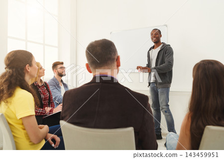 A speaker stands in front of a group during a workshop. Participants sit in chairs, listening and engaging. The room has large windows and a whiteboard. A speaker stands in front of a group during a workshop. Participants sit in chairs, listening and engaging. The room has large windows and a whiteboard. 143459951
