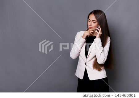 A woman stands in a suit while holding a phone to her ear. She appears thoughtful as she looks away. The background is gray and plain, focusing attention on her expression. A woman stands in a suit while holding a phone to her ear. She appears thoughtful as she looks away. The background is gray and plain, focusing attention on her expression. 143460001